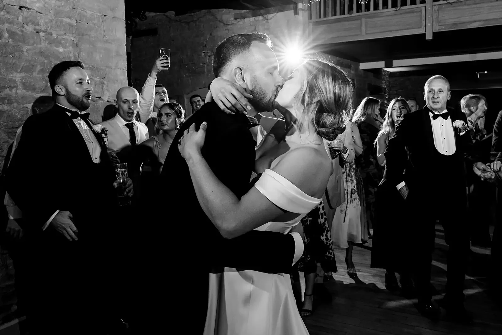 A black-and-white photo of a wedding reception showing a bride and groom sharing a kiss on the dance floor, surrounded by guests in formal attire, some singing and others watching.