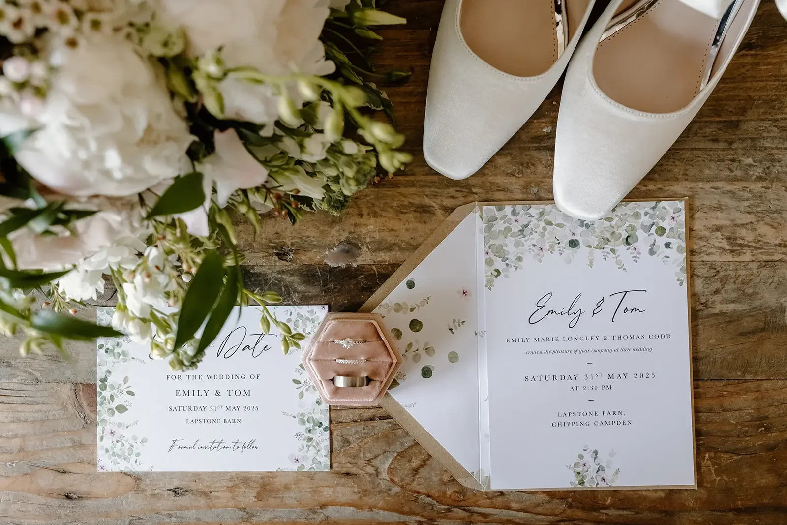 Wedding invitation and RSVP card for Emily and Tom, with a bouquet of white flowers and a pair of white high-heeled shoes on a wooden surface, featuring a pink ring box with rings inside.