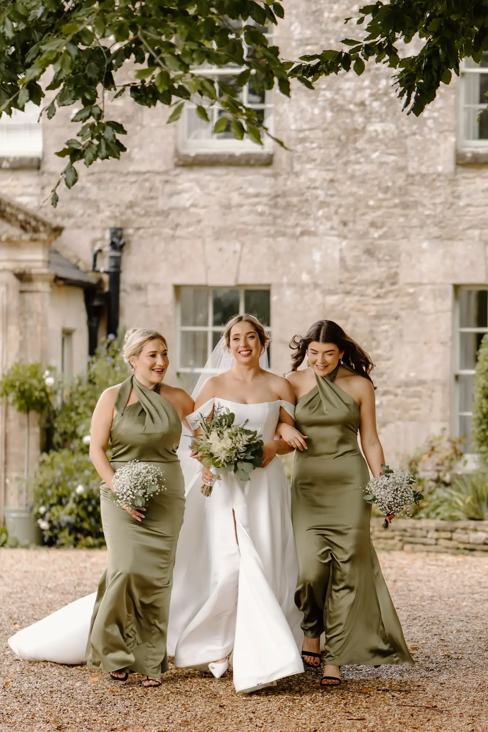A bride in a white wedding dress walking with two bridesmaids in olive green dresses holding bouquets, outside in front of an old stone building.