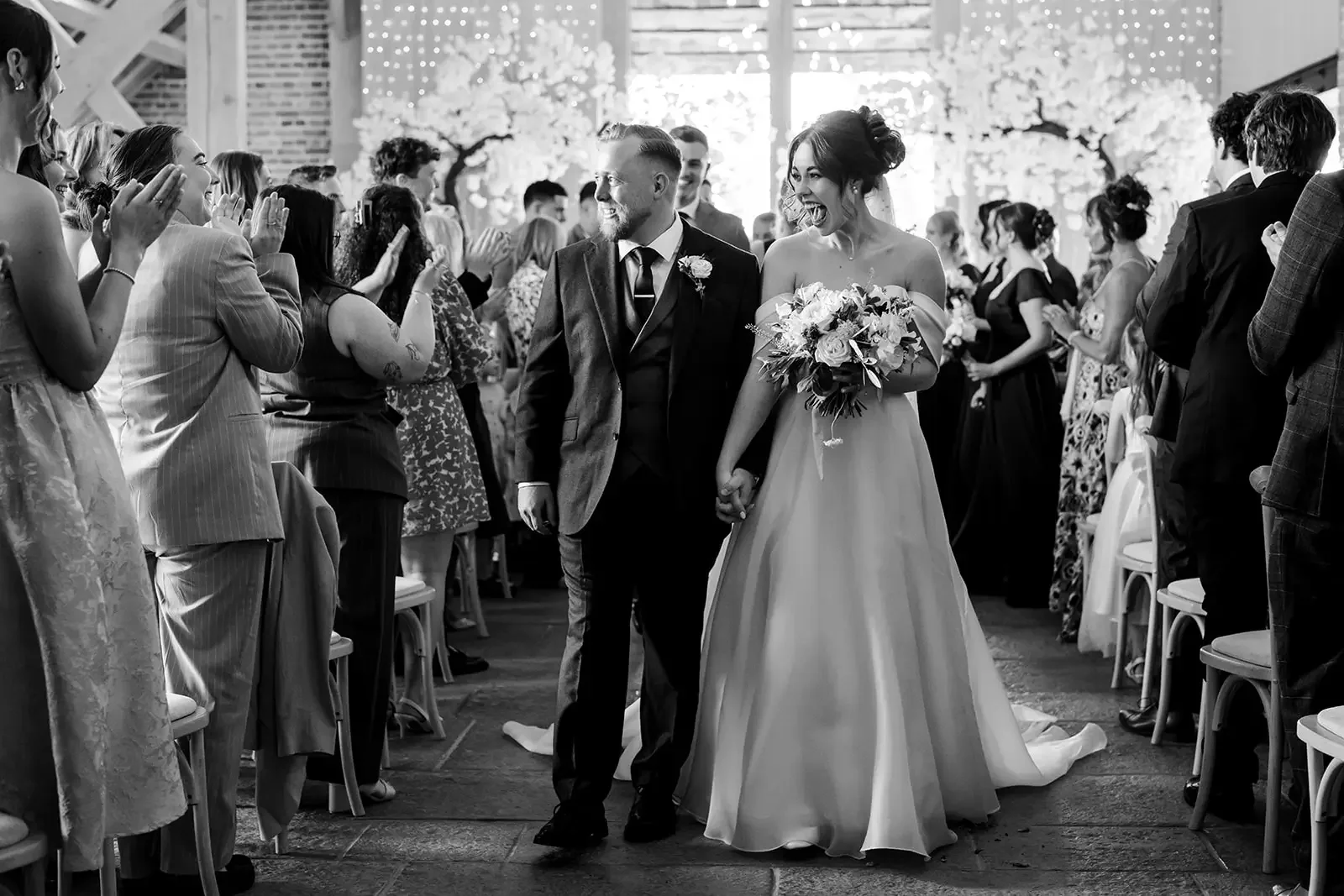 Black and white photograph of a wedding ceremony with a bride and groom holding hands and walking down the aisle, surrounded by guests who are clapping and smiling.