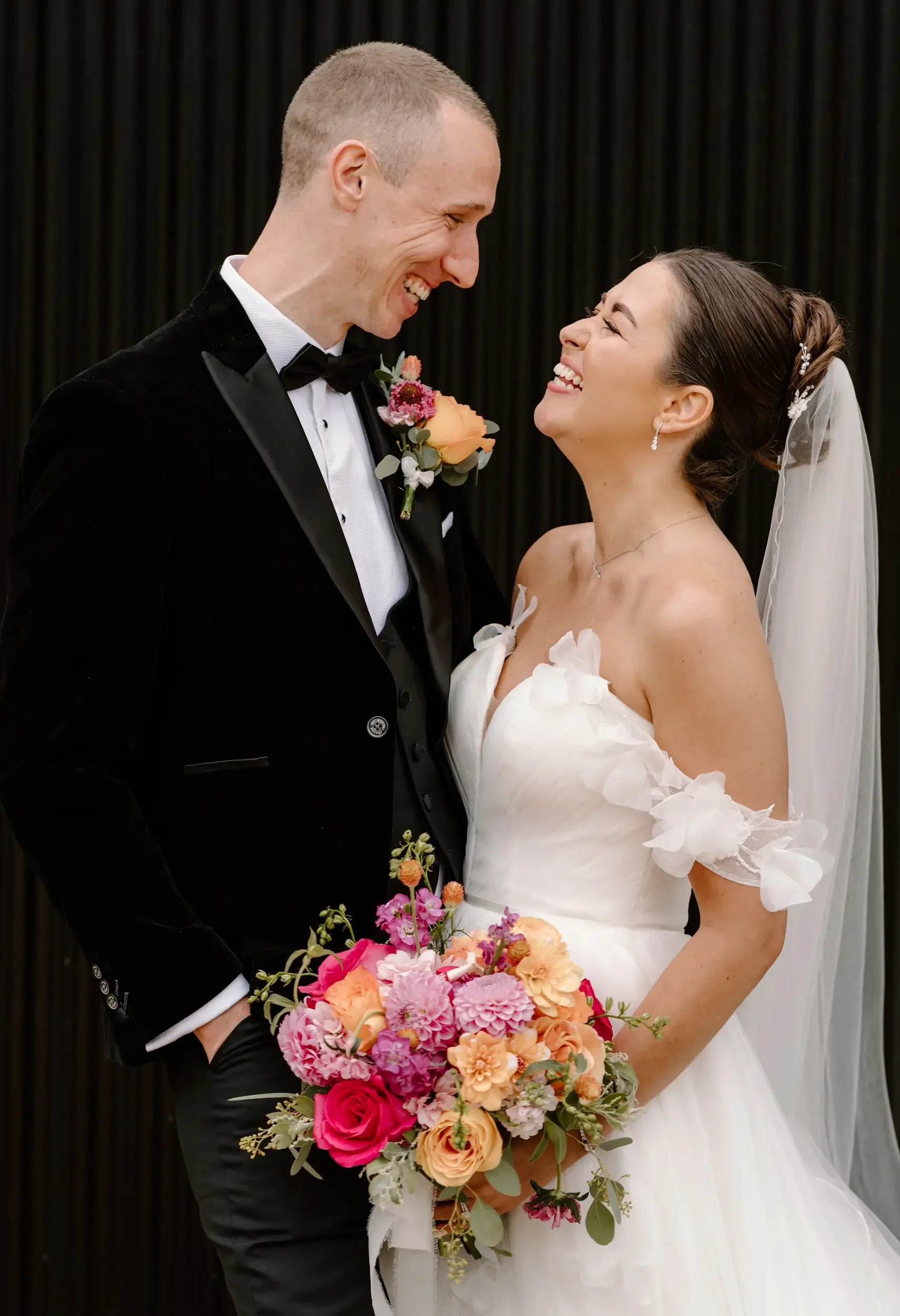 A bride and groom smiling and looking into each other's eyes, with the bride holding a colorful bouquet, against a dark backdrop.