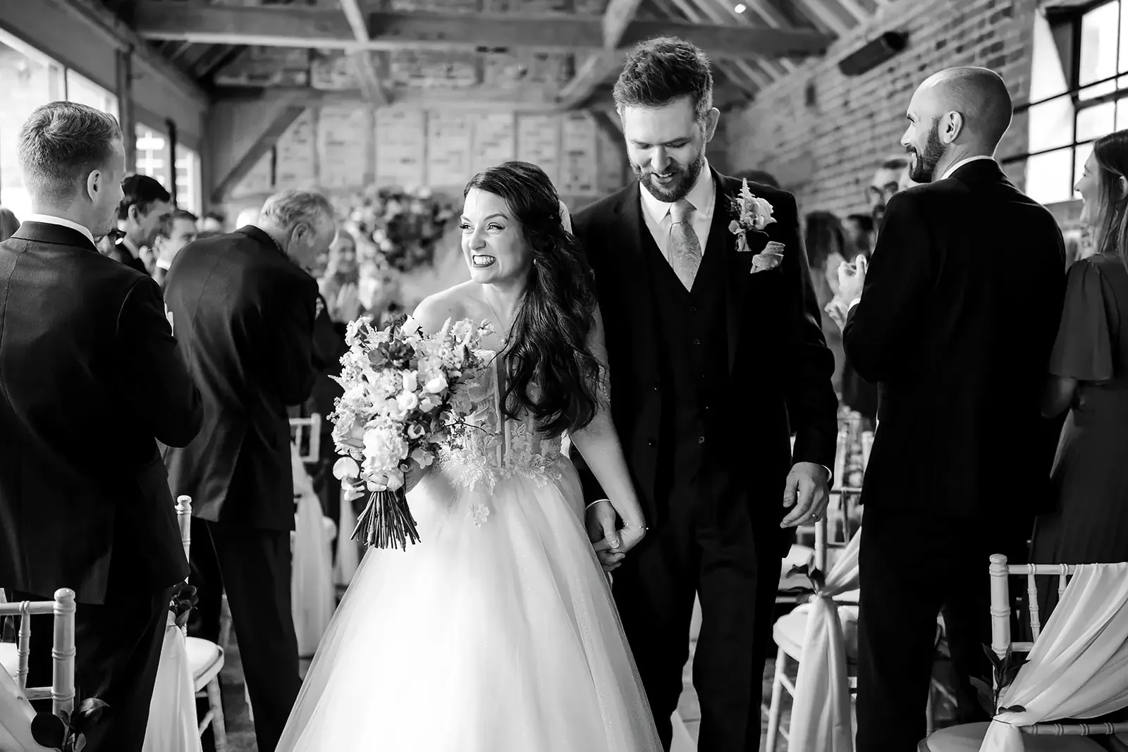 Black and white photo of a bride and groom holding hands and smiling, walking down the aisle during their wedding ceremony, surrounded by guests.