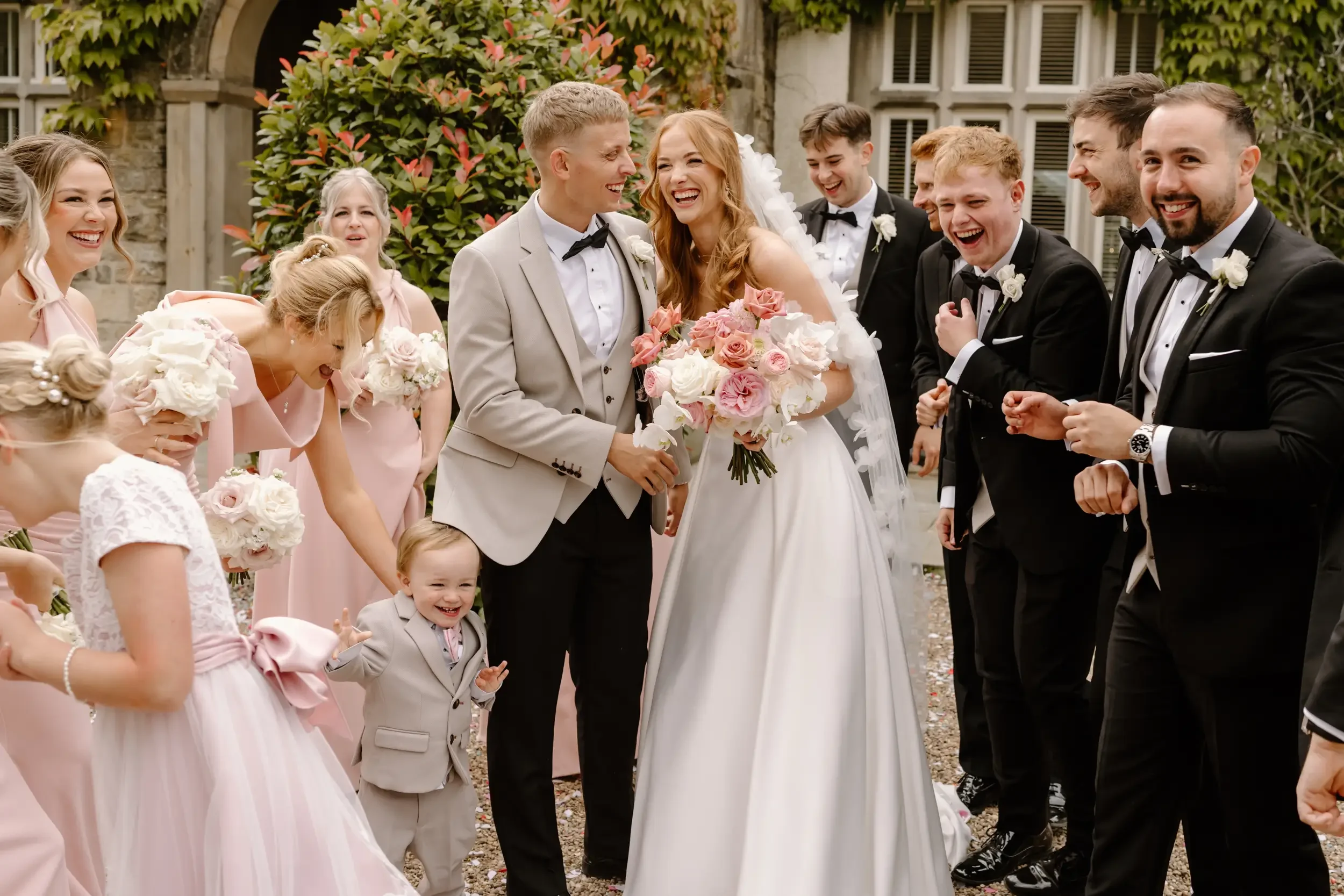 Bride and groom smiling, surrounded by wedding party and guests, outdoors in front of a stone building with greenery.