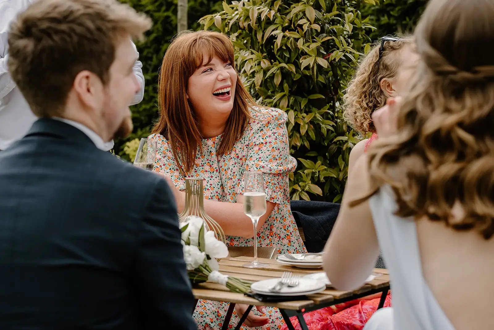 A woman with red hair is laughing and sitting at a table outdoors with a group of people during a social gathering, with some of the others partially visible around her.