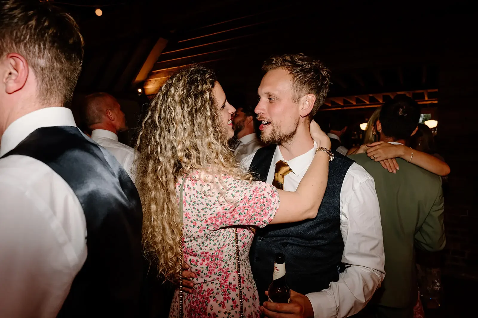 A couple dancing closely, a woman with curly hair and a man with short hair, in a crowded indoor setting with warm lighting, some other people dancing in the background.