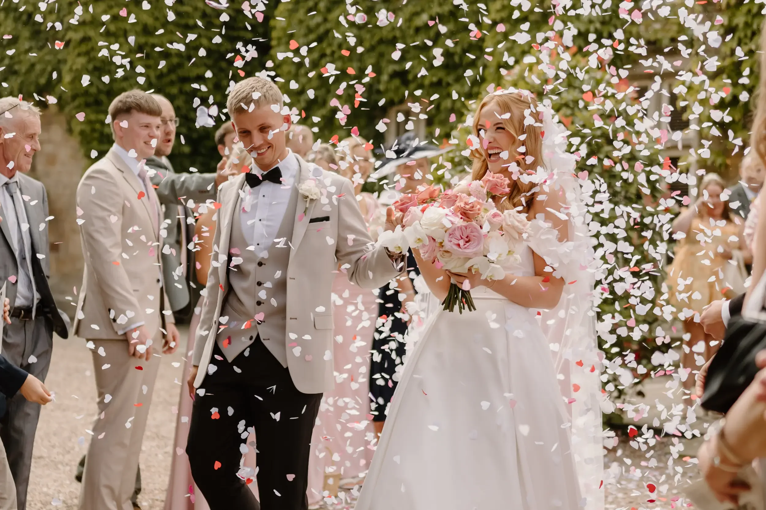 Bride and groom smiling, surrounded by guests, as confetti falls at outdoor wedding celebration.
