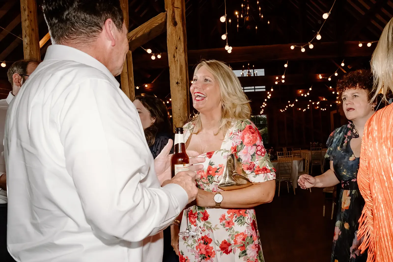 People talking and laughing at an indoor social event with string lights hanging from the ceiling, rustic wooden beams and tables in the background.