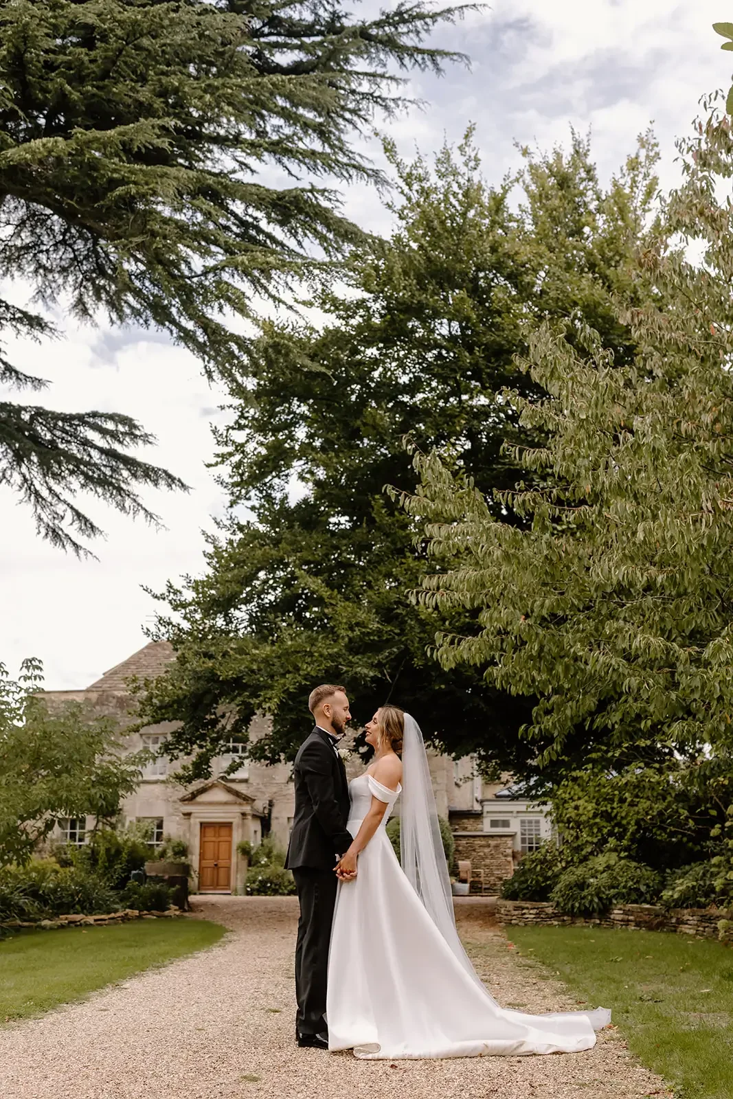 A bride and groom standing hand in hand on a gravel path in front of a large house, surrounded by trees and greenery, during daytime.