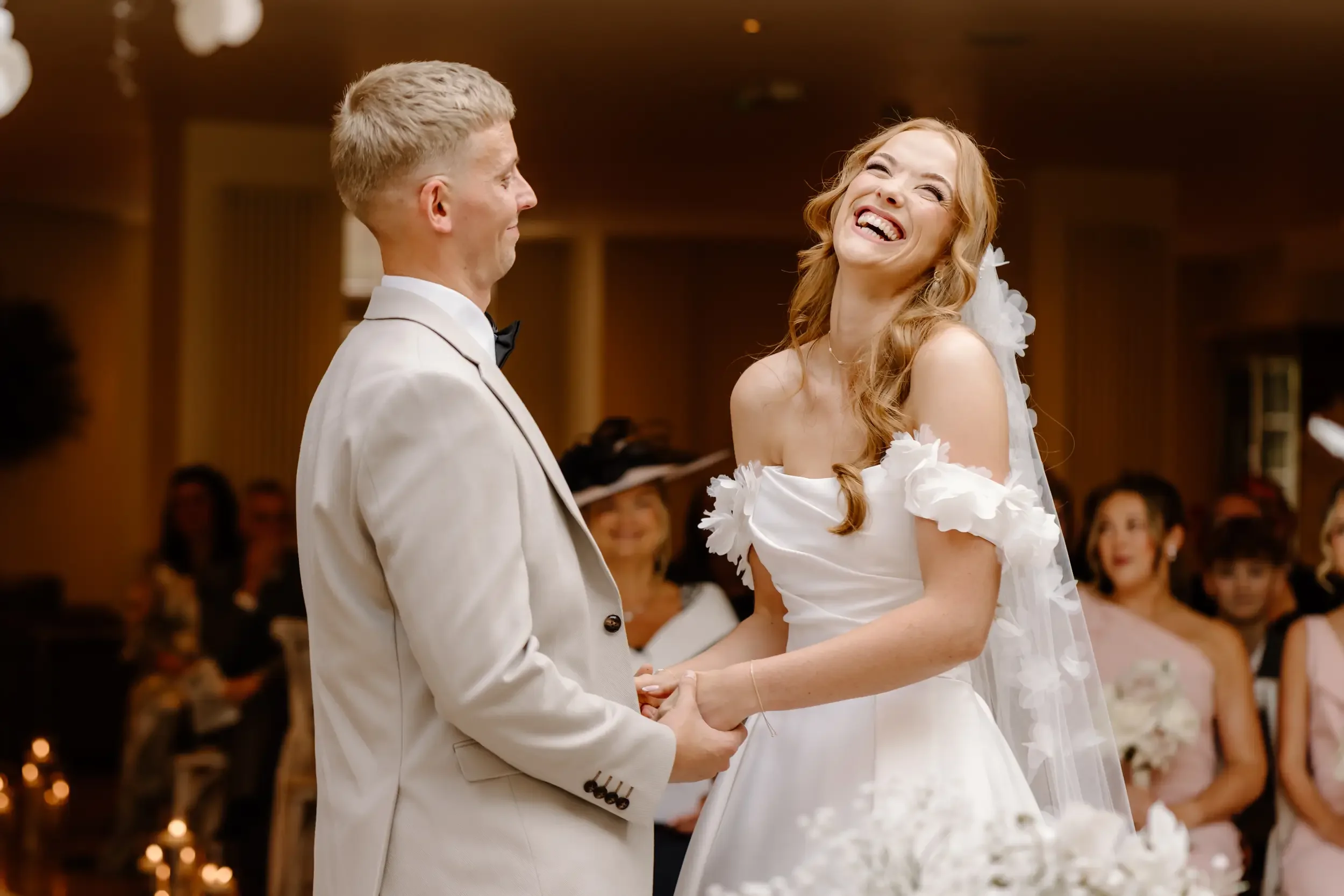 A bride and groom sharing a moment during their wedding ceremony, holding hands and smiling at each other in a warmly lit room.