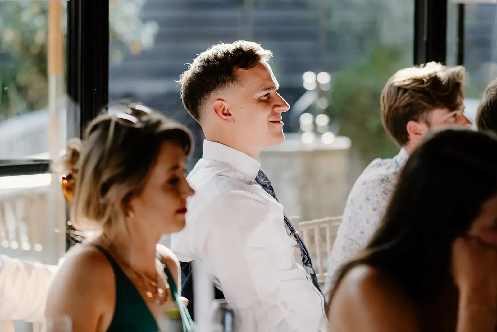 People dressed in formal attire sitting indoors during daytime, with sunlight streaming through large windows in the background.