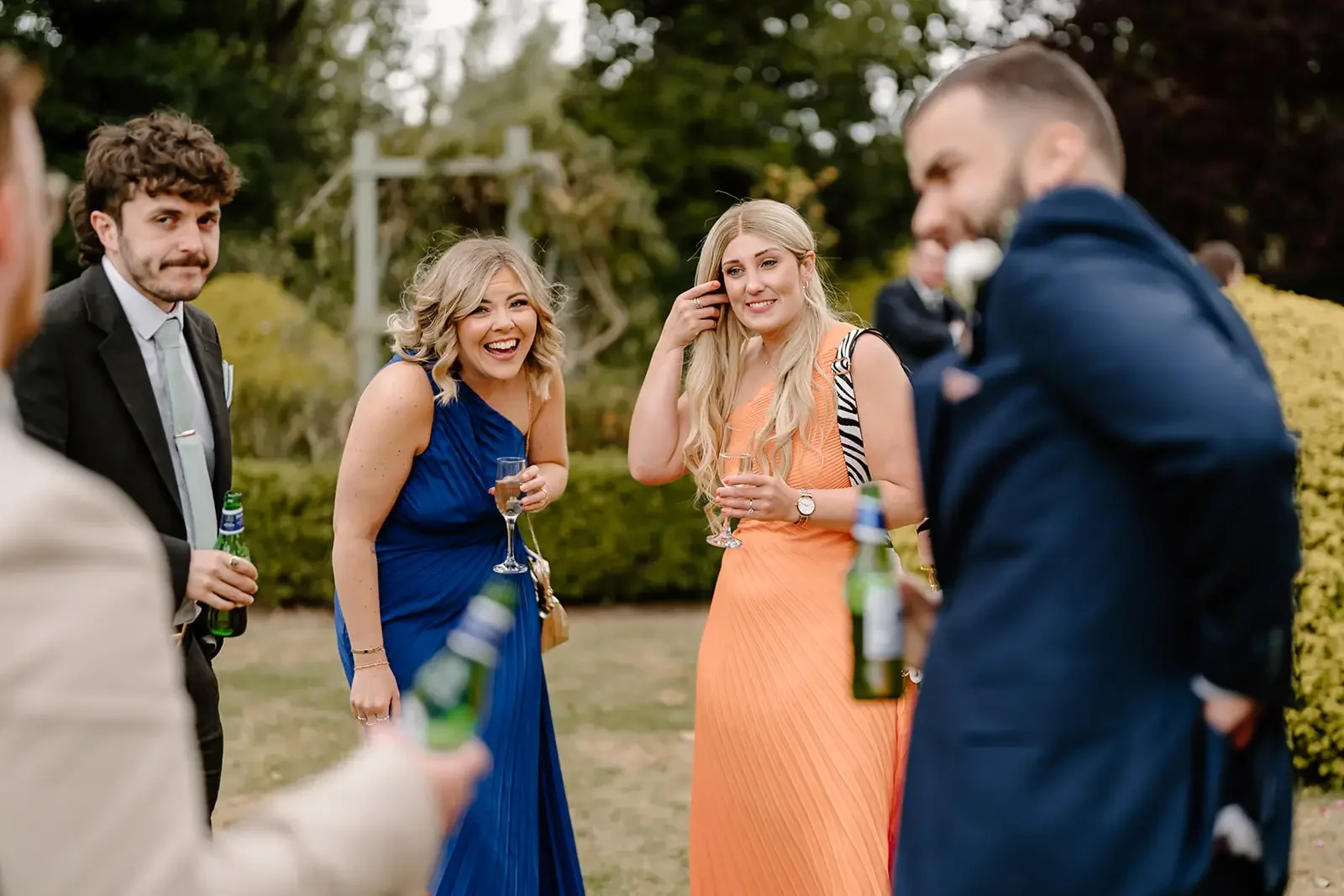 Group of people at an outdoor social gathering, holding drinks and talking, with trees and a garden in the background.