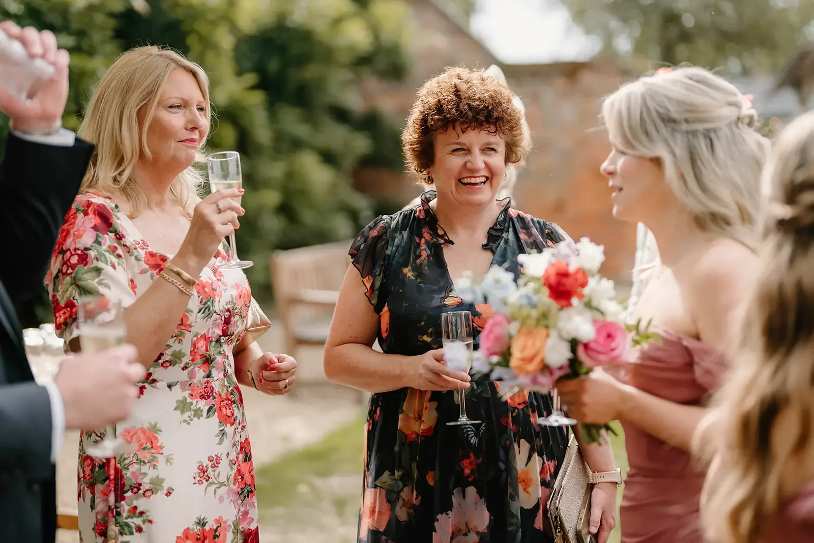 Group of women at an outdoor celebration, smiling and holding glasses of champagne, with a bouquet of flowers in the foreground.