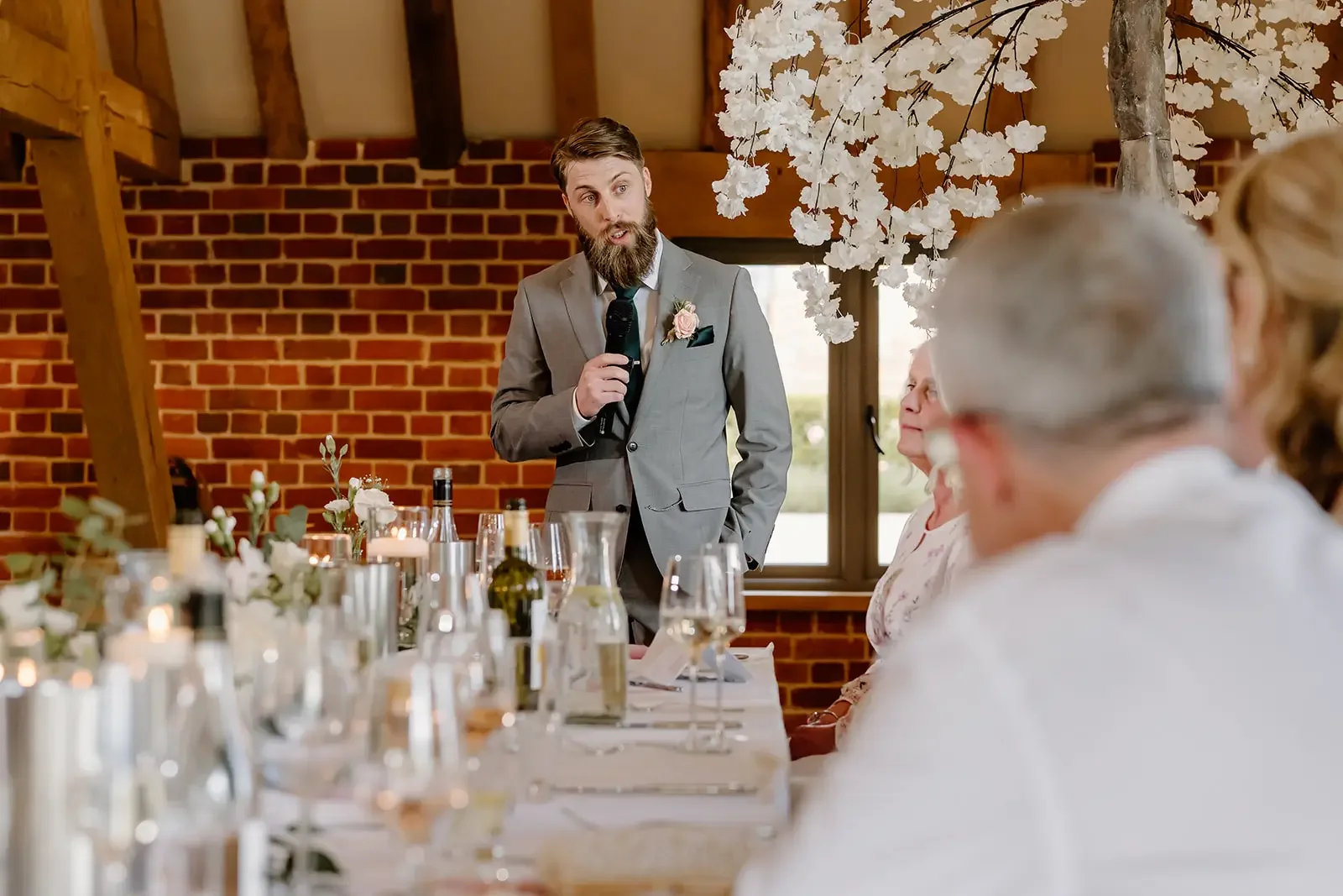 A man in a gray suit giving a speech at a wedding reception, with guests seated at a decorated table, in a rustic venue with brick walls and wooden beams.