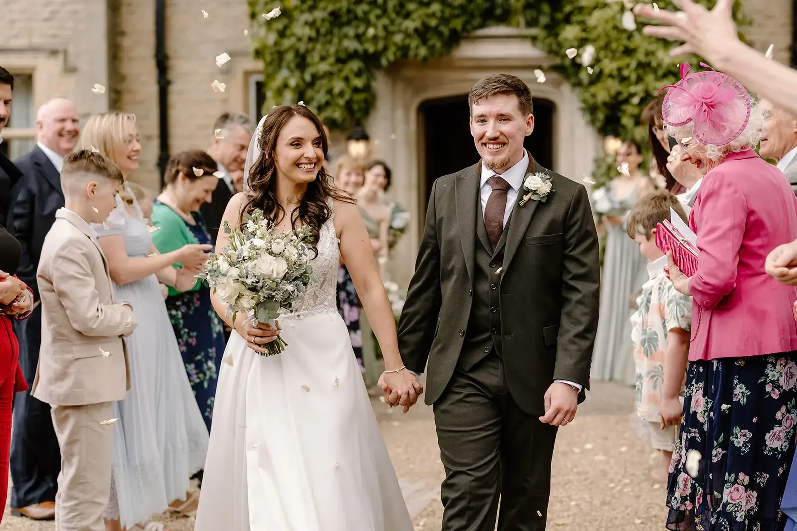 Wedding couple holding hands, smiling, walking outside surrounded by guests, woman in white wedding dress holding a bouquet of white flowers, man in dark suit, guests dressed in formal attire, some with hats, celebrating with confetti.
