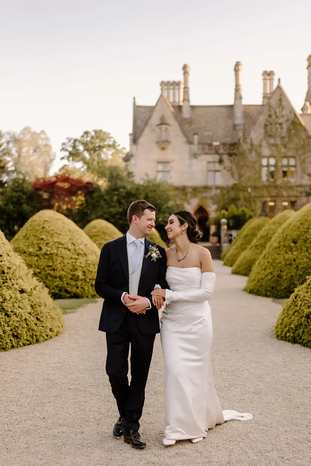 A newlywed couple walking arm-in-arm on a garden path, with a historic stone castle and lush greenery in the background during sunset.