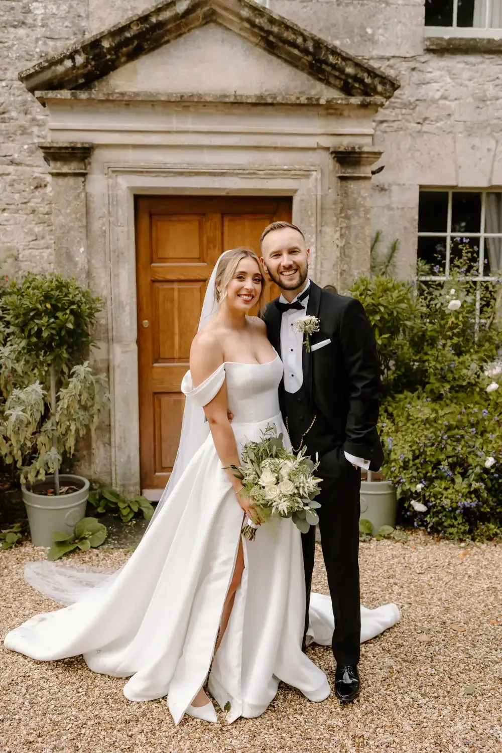 A bride and groom standing outside a stone building, smiling. The bride is holding a bouquet and wearing a white wedding dress with a slit, and the groom is in a black tuxedo with a bow tie. There is greenery and potted plants around them.