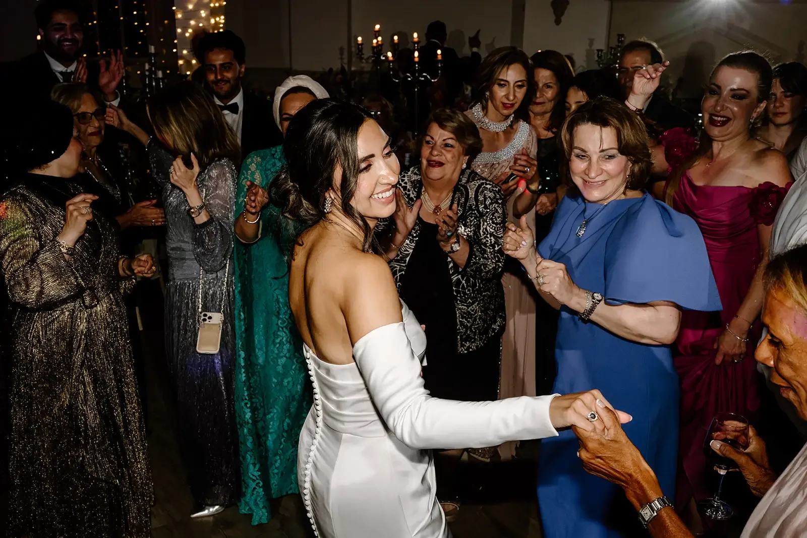 A bride dancing with an older woman at a wedding reception, surrounded by guests in formal attire, with warm lighting and elegant decorations in the background.