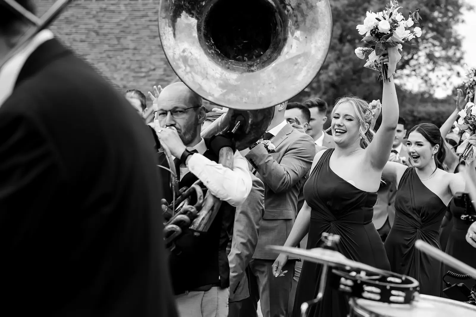 A black-and-white photo of a group of people at a celebration or wedding, with some playing musical instruments and others dancing and smiling, holding bouquets of flowers, outdoors.