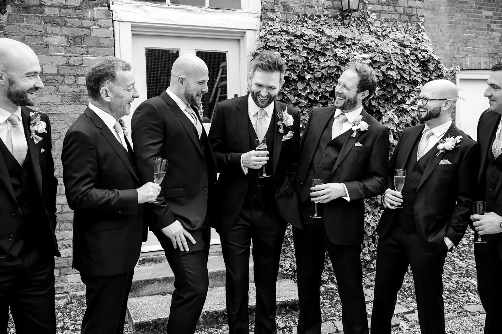 Group of men in suits at a wedding celebration, holding glasses of champagne, standing outdoors against a brick wall with ivy.