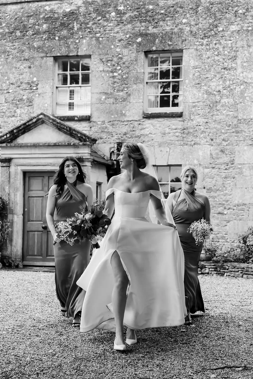 Black and white photo of a bride in a wedding dress walking outdoors with two bridesmaids holding bouquets, in front of a stone building with windows.