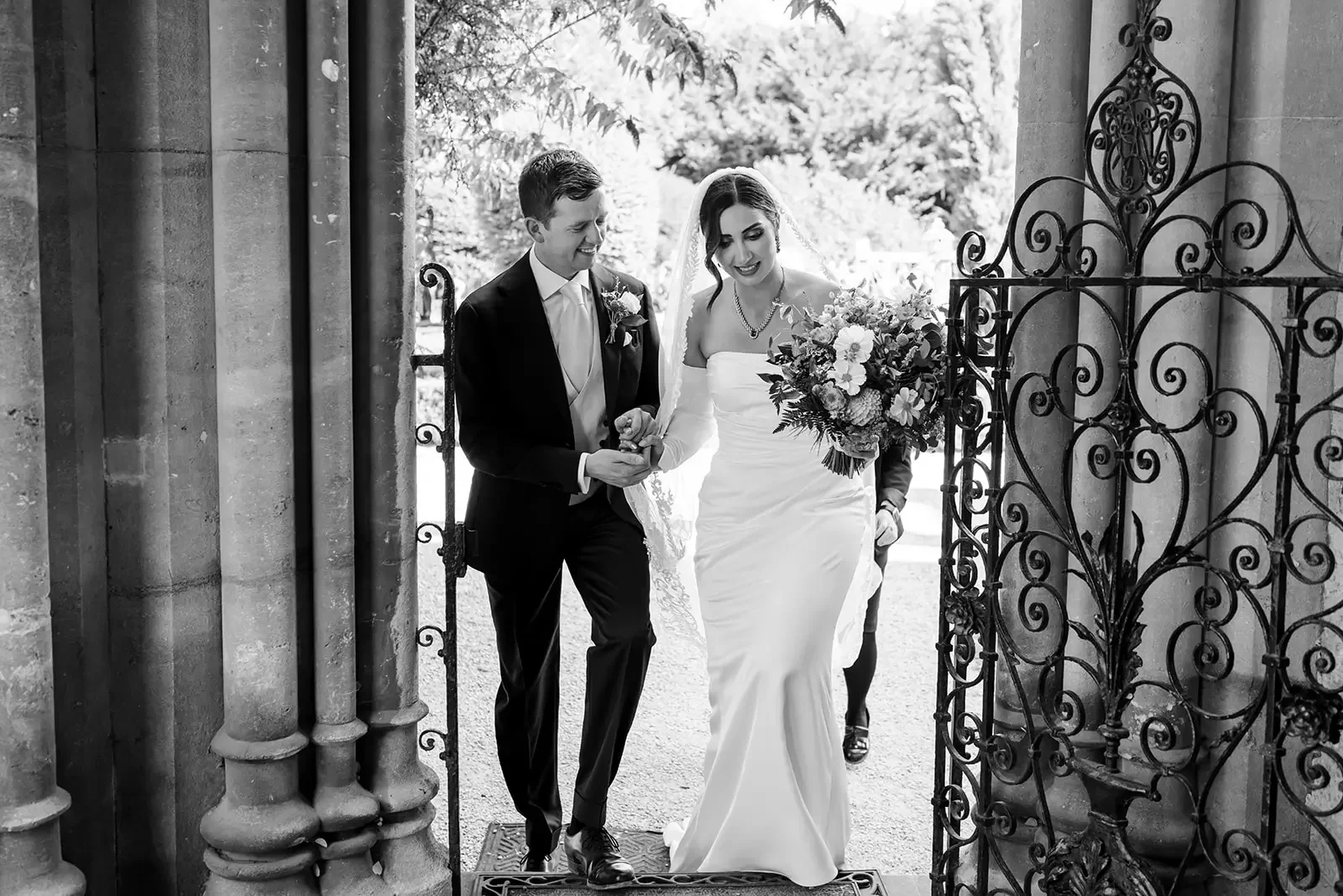 A black and white photo of a bride and groom walking through a wrought iron gate, with a bride holding a bouquet of flowers, as they smile and look happy.