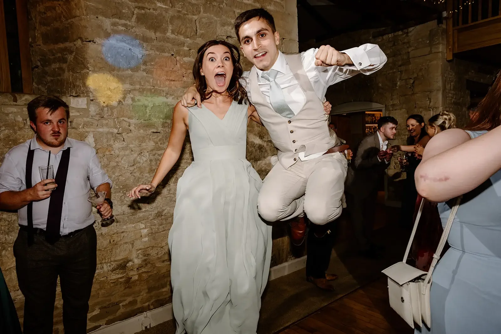 A woman in a white dress and a man in a white vest and shirt are jumping and celebrating at a wedding reception with other guests in the background.
