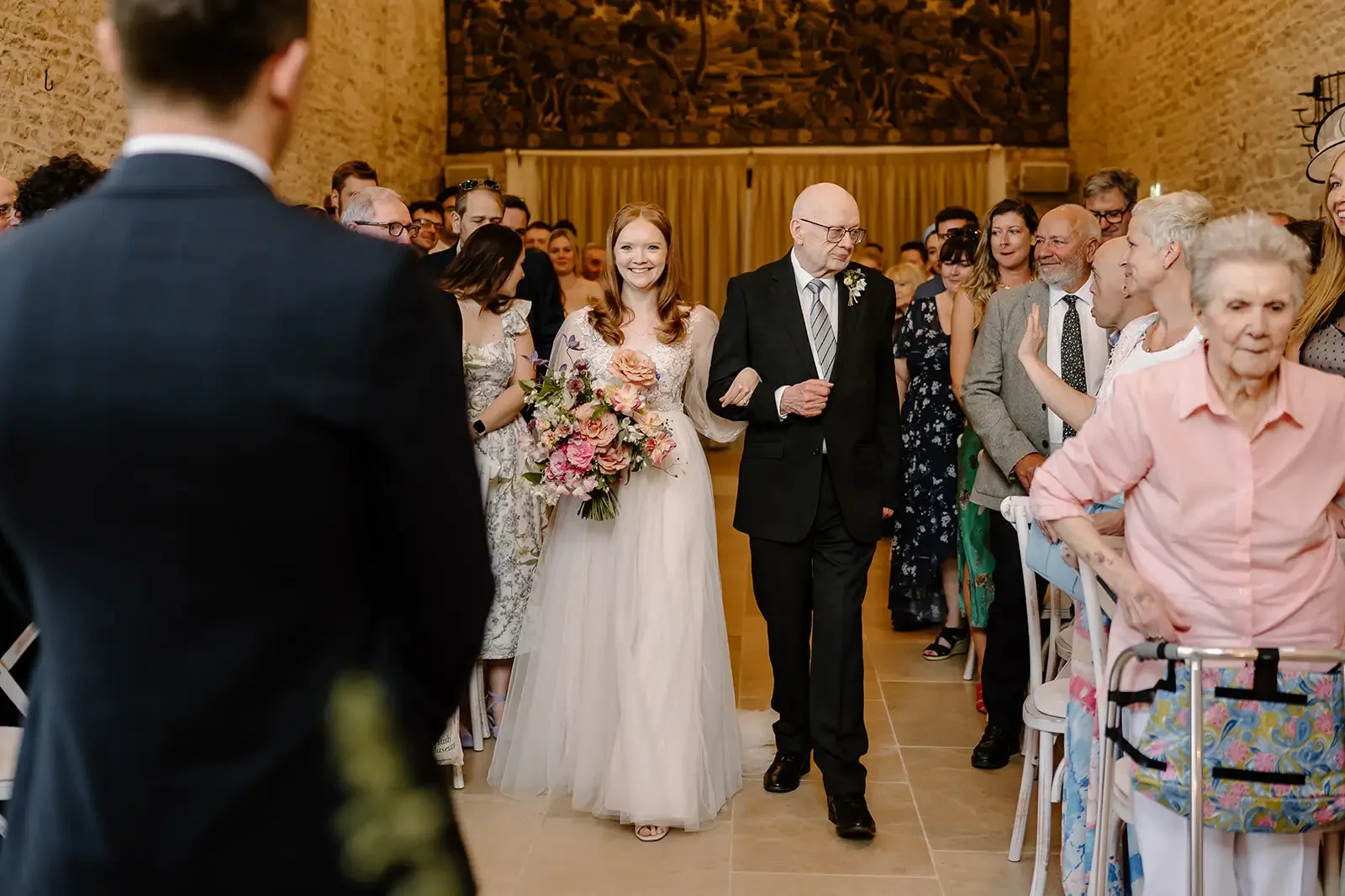 A bride walking down the aisle with her father at a wedding ceremony, surrounded by seated guests in a warmly lit indoor venue.
