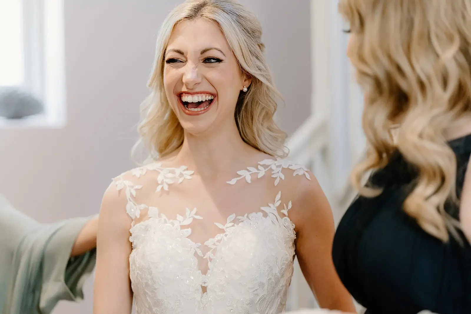 A woman in a white wedding dress with floral lace details on the shoulders is laughing with her eyes closed and mouth open, during a joyful moment.