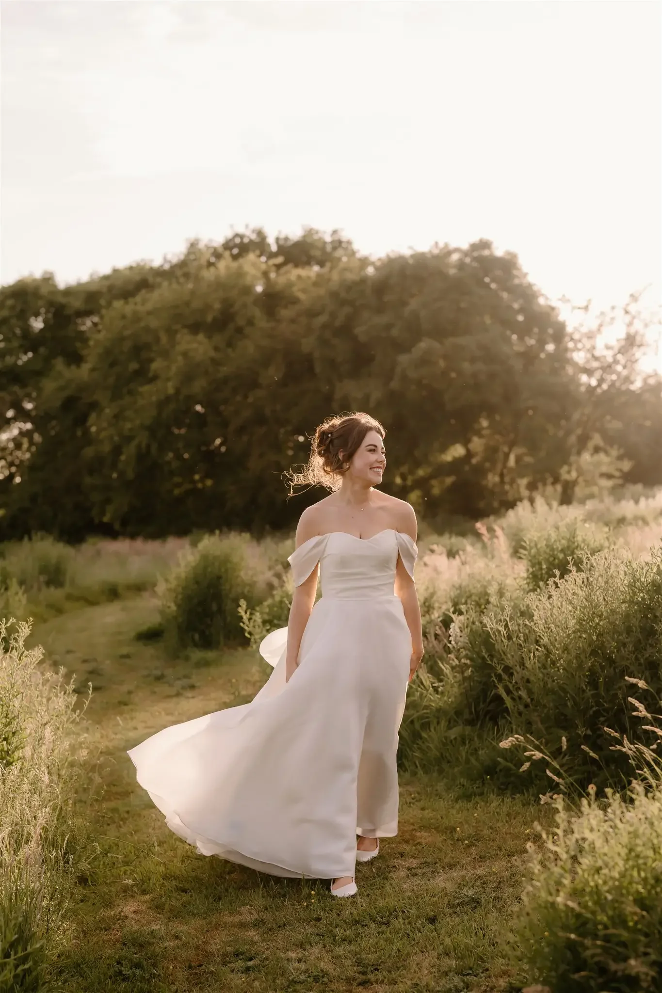 A woman in a white off-the-shoulder wedding dress walking on a grassy path in a field during sunset.