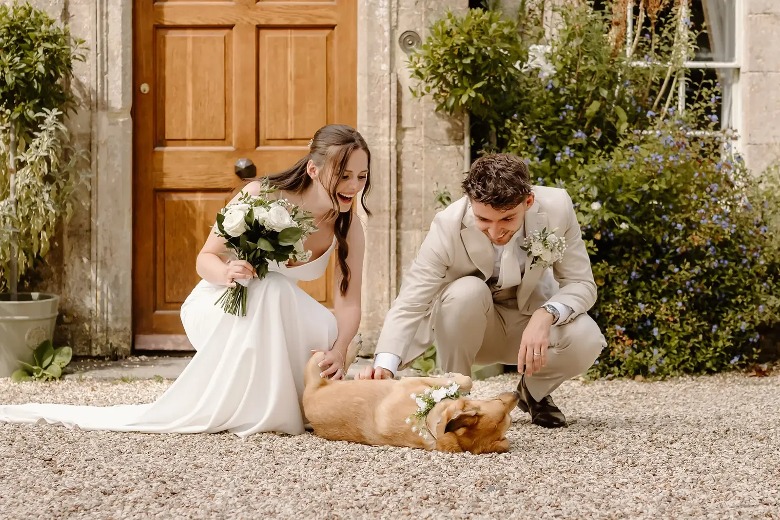 A bride and groom smiling and playing with a dog in front of a house door, with the bride holding a bouquet and both wearing wedding attire.