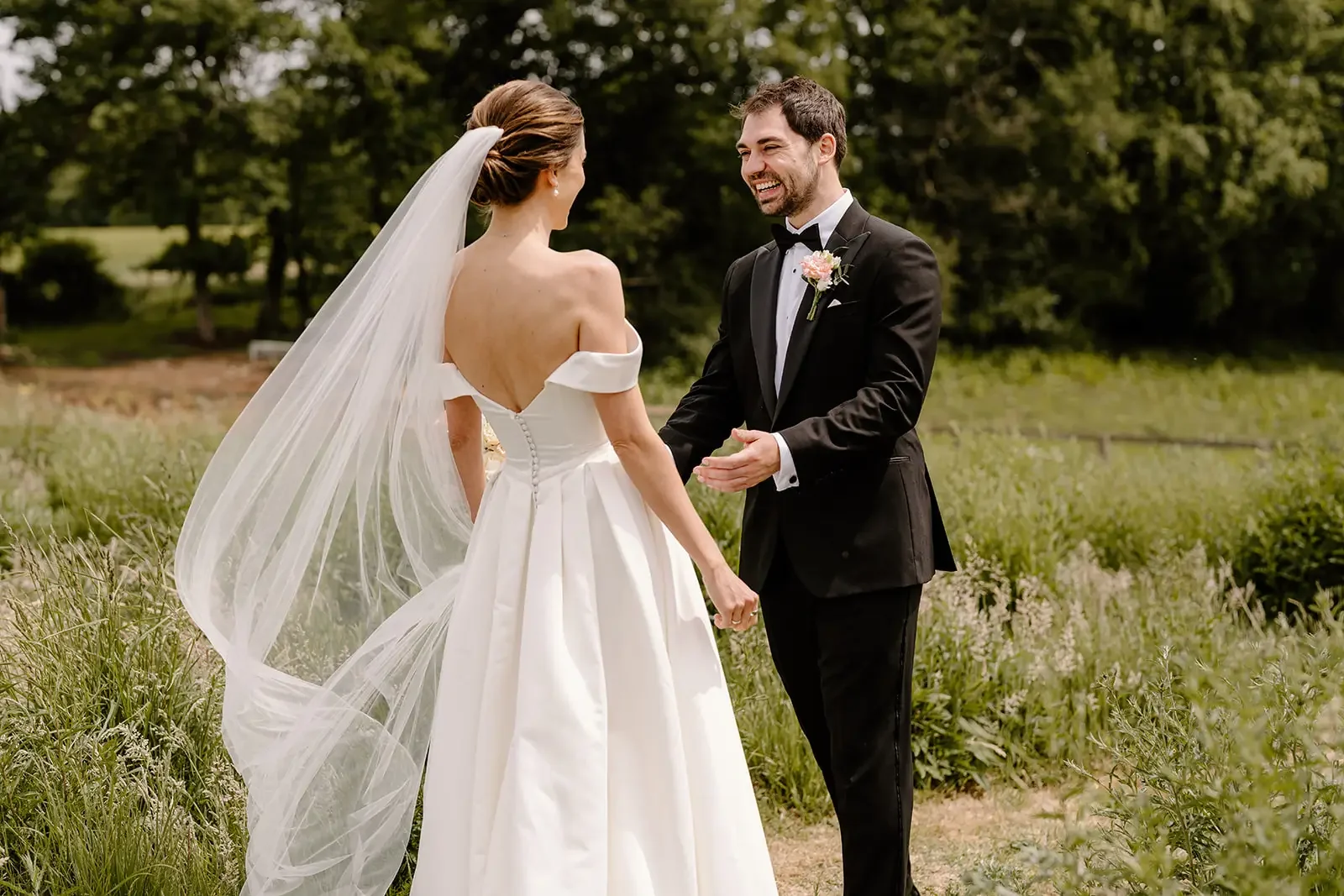 A bride and groom holding hands outdoors on their wedding day, with trees and greenery in the background.