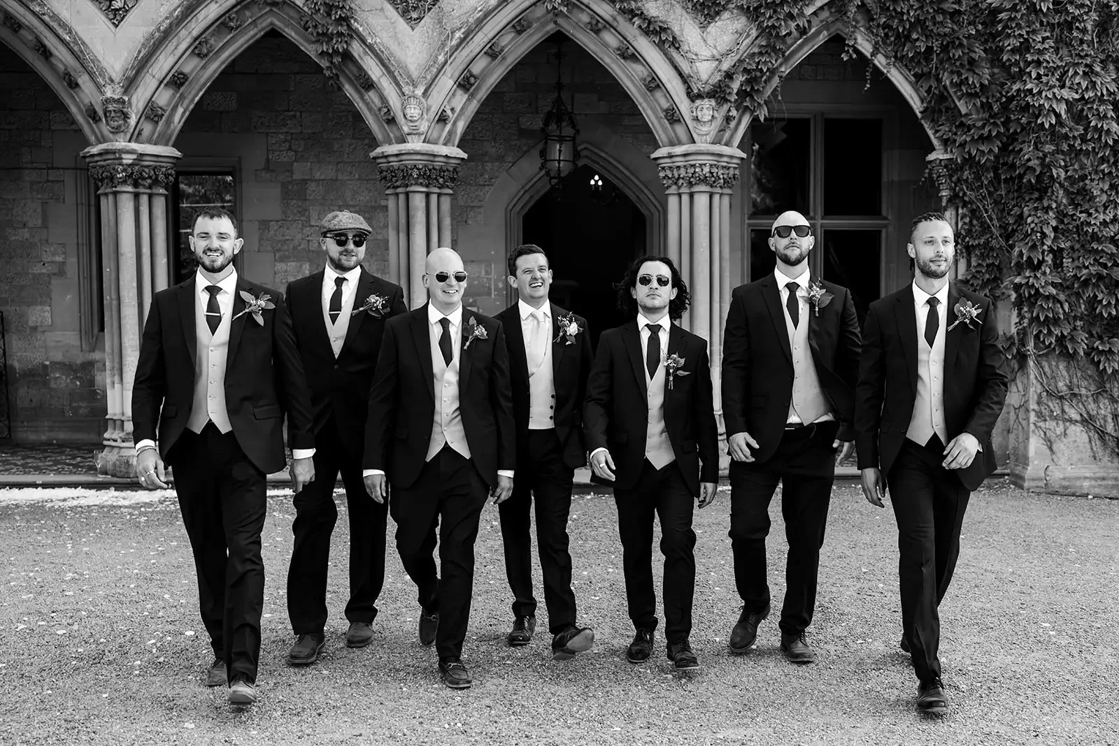 Black and white photo of seven men in suits walking confidently in front of a historic building with Gothic arches and ivy growing on the walls.