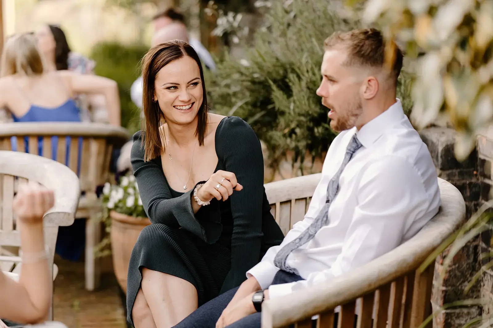 A woman and a man are sitting on a bench outdoors, smiling and engaging in conversation, with other people sitting at tables in the background.