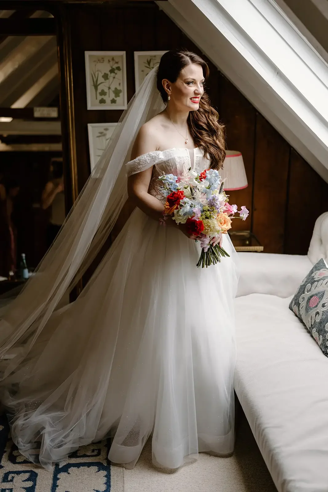 A smiling bride in an off-shoulder wedding gown with a long veil, holding a colorful bouquet, standing indoors near a white couch and a dark wooden wall with framed botanical prints.