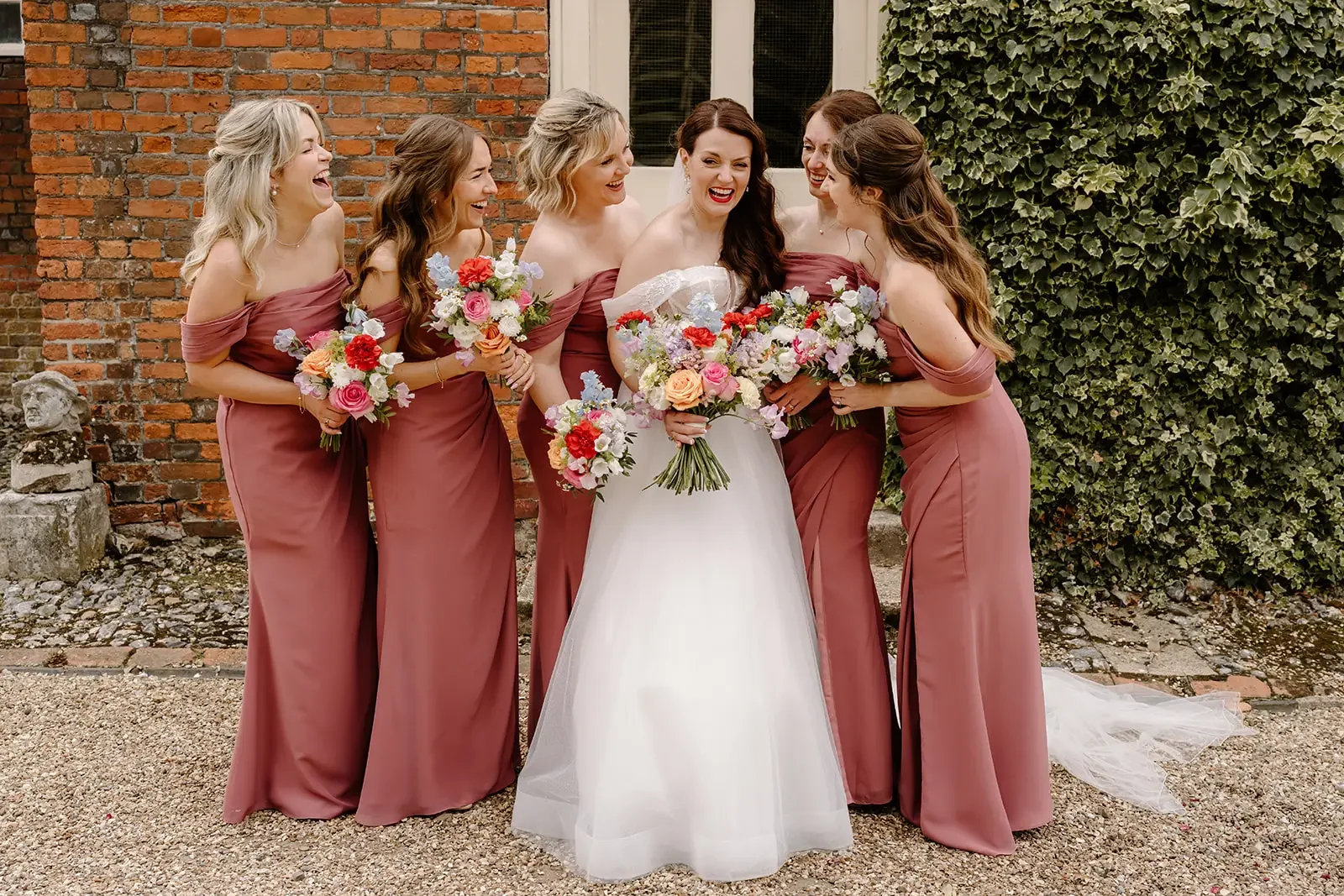 Bride in white wedding dress with five bridesmaids in mauve dresses, all holding colorful bouquets, standing outside against a brick and ivy wall.