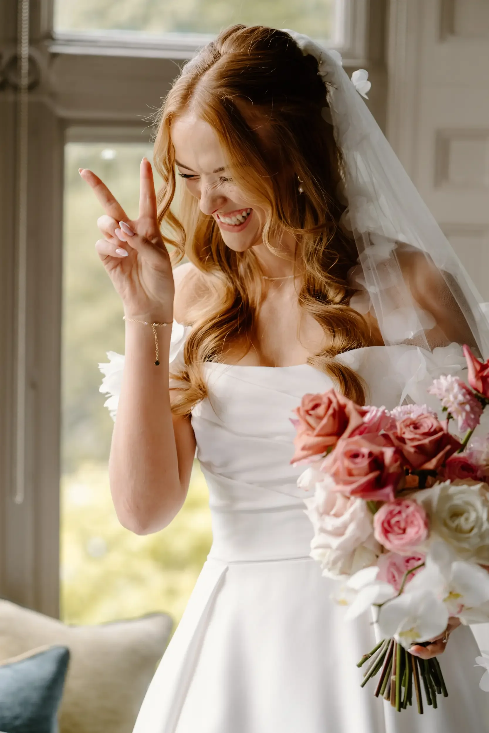 A smiling bride with red hair making a peace sign with her fingers, holding a bouquet of pink and white flowers, wearing a white wedding dress and veil, standing near a window.