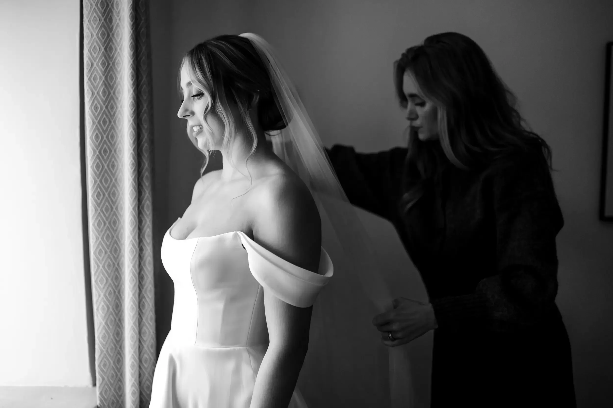 A bride in a wedding dress with off-the-shoulder sleeves is being helped with her veil by a woman in a dark outfit, in a softly lit room.