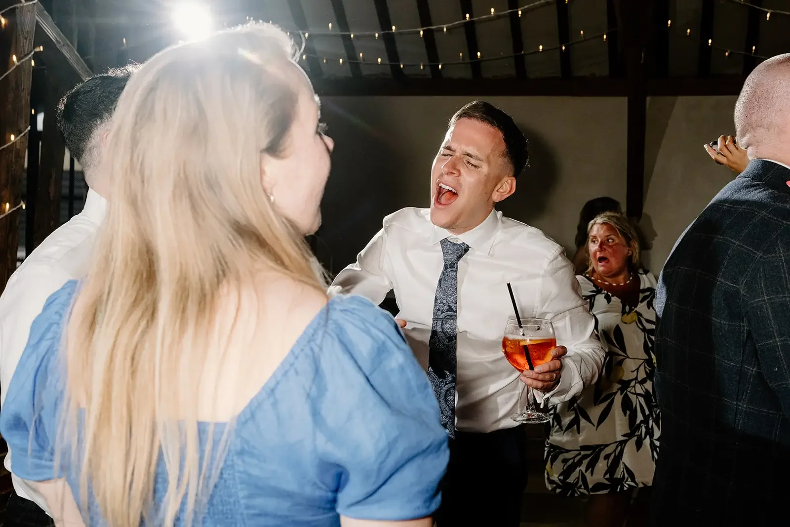 People socializing at a party or celebration, with a man singing and holding a drink, surrounded by women and others in a decorated indoor setting.