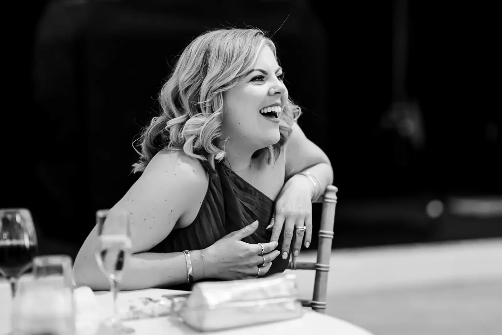 A woman with curly blonde hair laughing, sitting at a table, in a black dress, in a black and white photo.