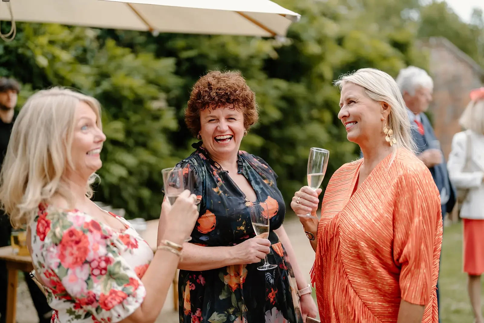 Three women smiling and talking while holding glasses of champagne at an outdoor gathering with greenery and other guests in the background.