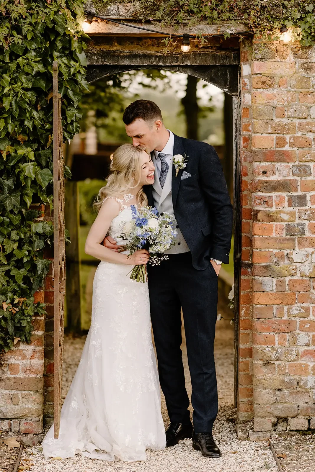 A bride and groom sharing a joyful moment under a rustic brick archway. The bride is holding a bouquet of blue and white flowers, and both are smiling and close together. The groom is kissing the bride's forehead.