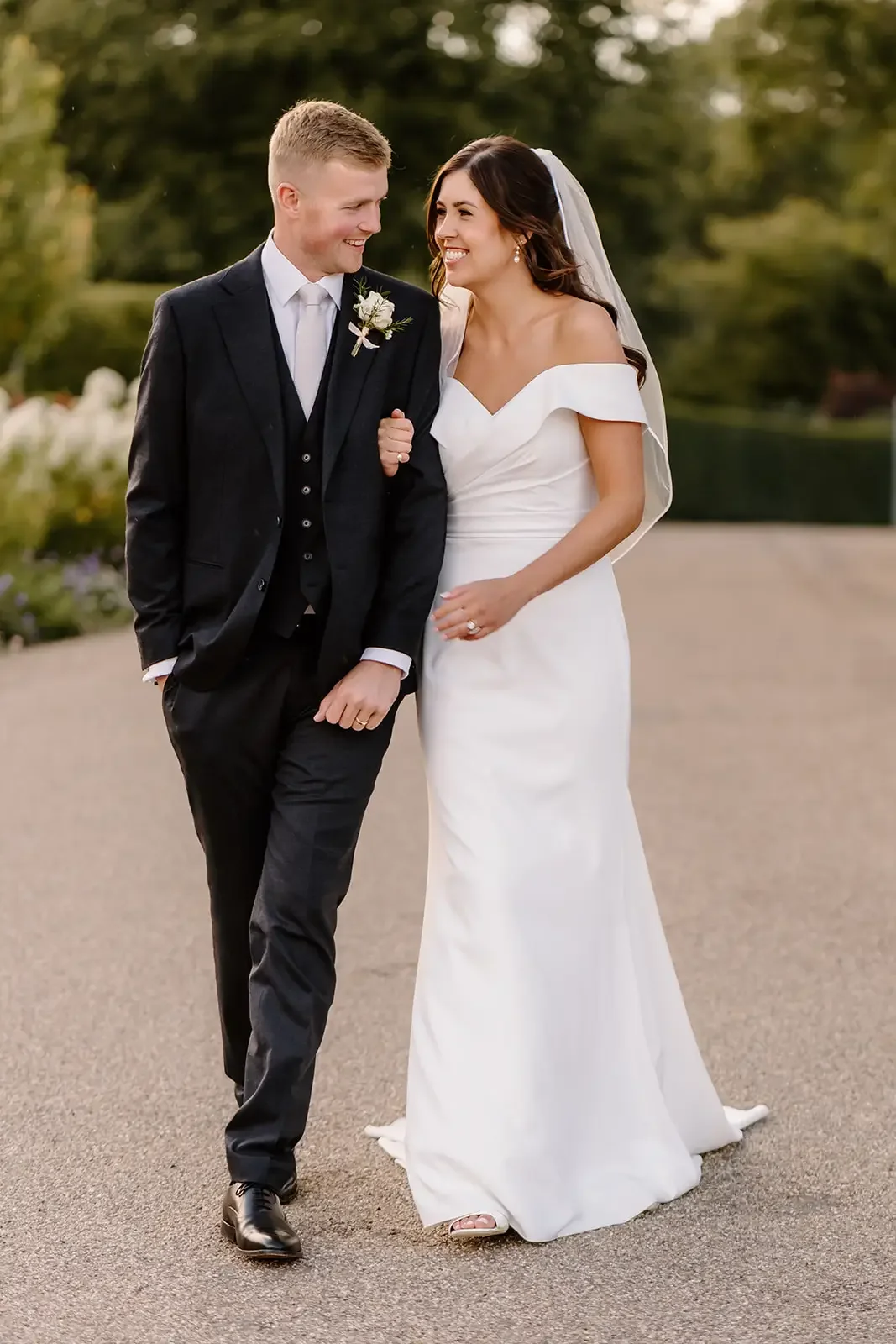 A newlywed couple walking outdoors, smiling at each other, with the bride wearing a white off-the-shoulder wedding dress and veil, and the groom in a black suit with a white shirt and boutonniere.
