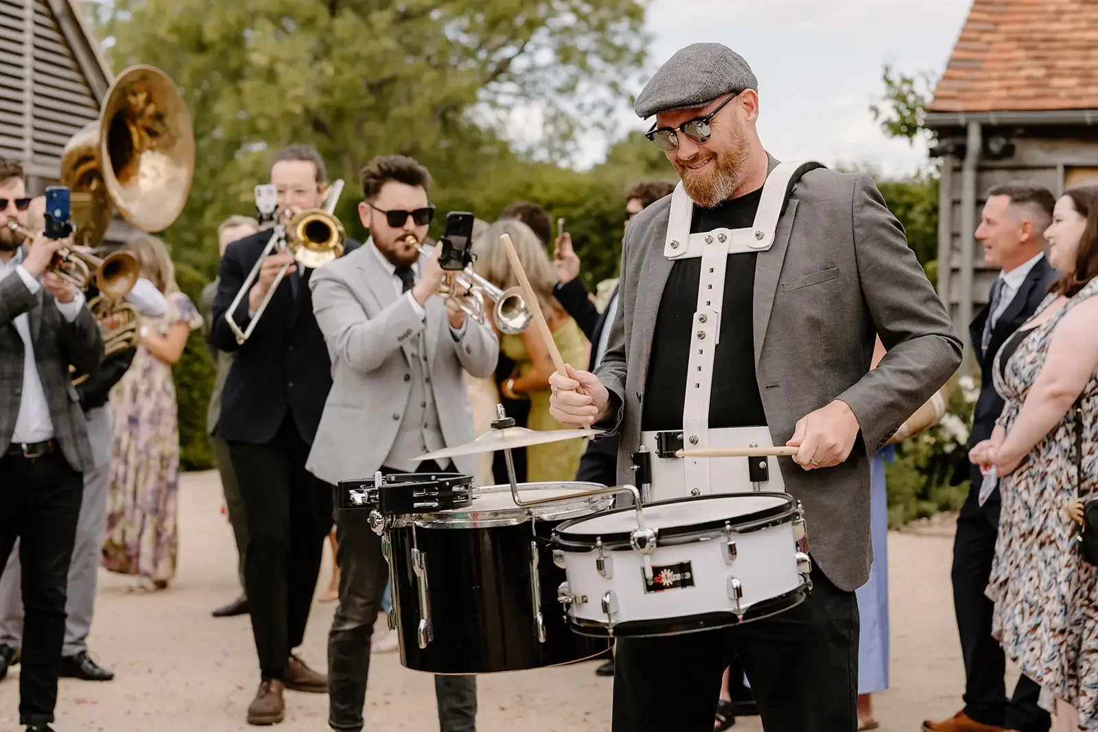 Man in gray blazer and flat cap playing a marching snare drum at outdoor event, surrounded by musicians with brass instruments and spectators in the background.