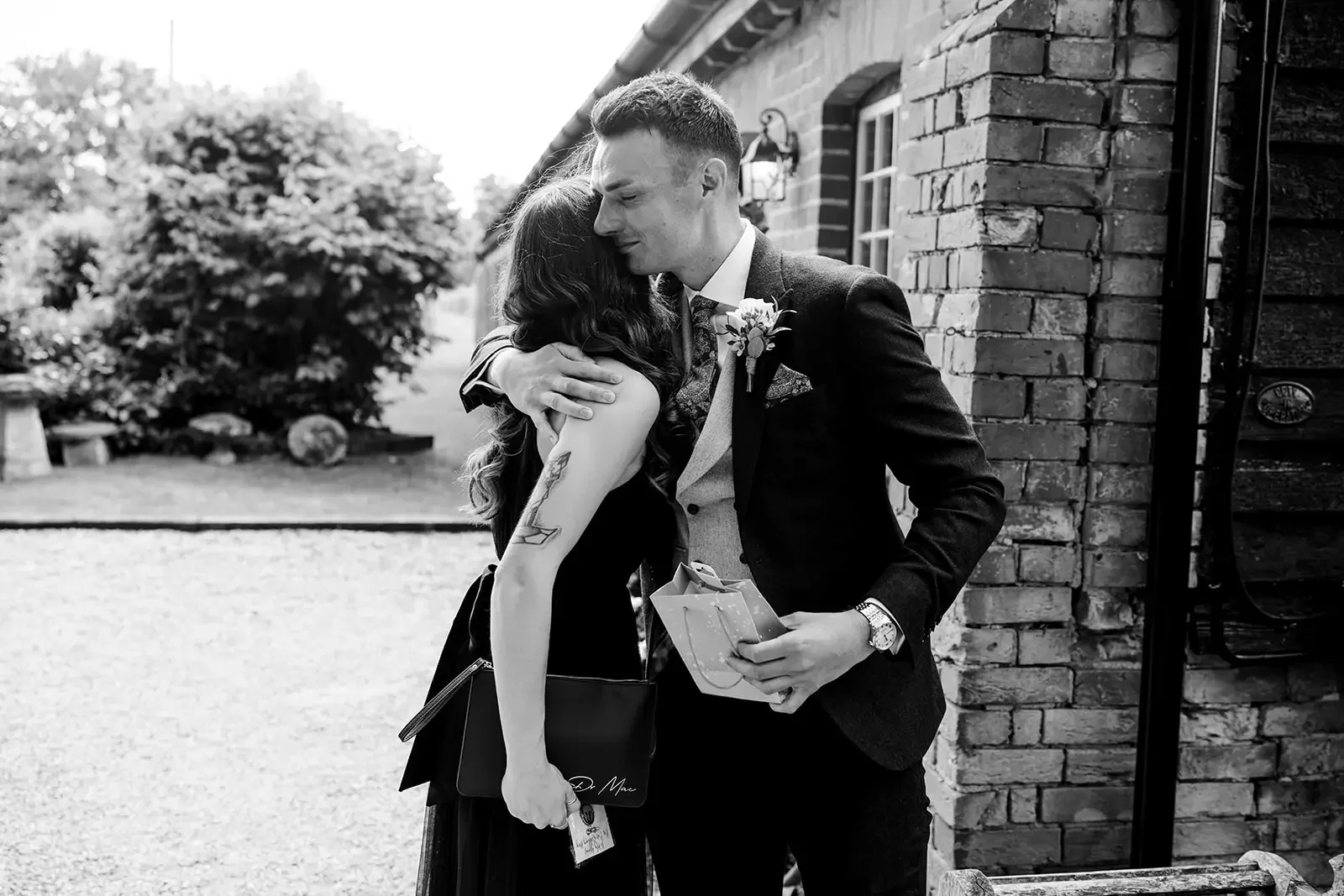A man in a suit hugging a woman in black dress outside a brick building, holding a gift and a bag.