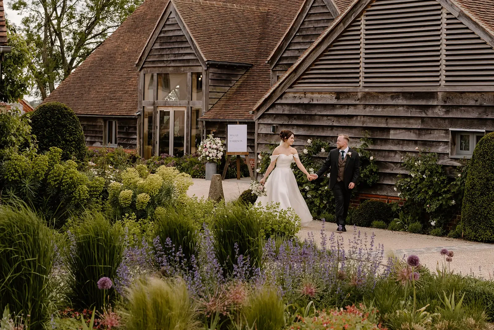 A bride and groom holding hands and walking outside a rustic building with wooden siding and a shingled roof, surrounded by lush green plants and flowers.