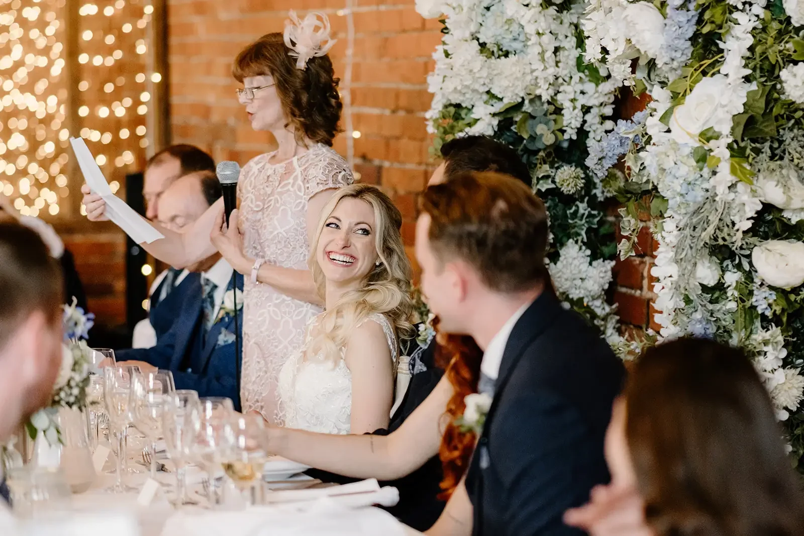 A woman in a lace dress giving a speech at a wedding reception, standing by a seated bride and groom, with guests seated at a long table and floral decorations in the background.