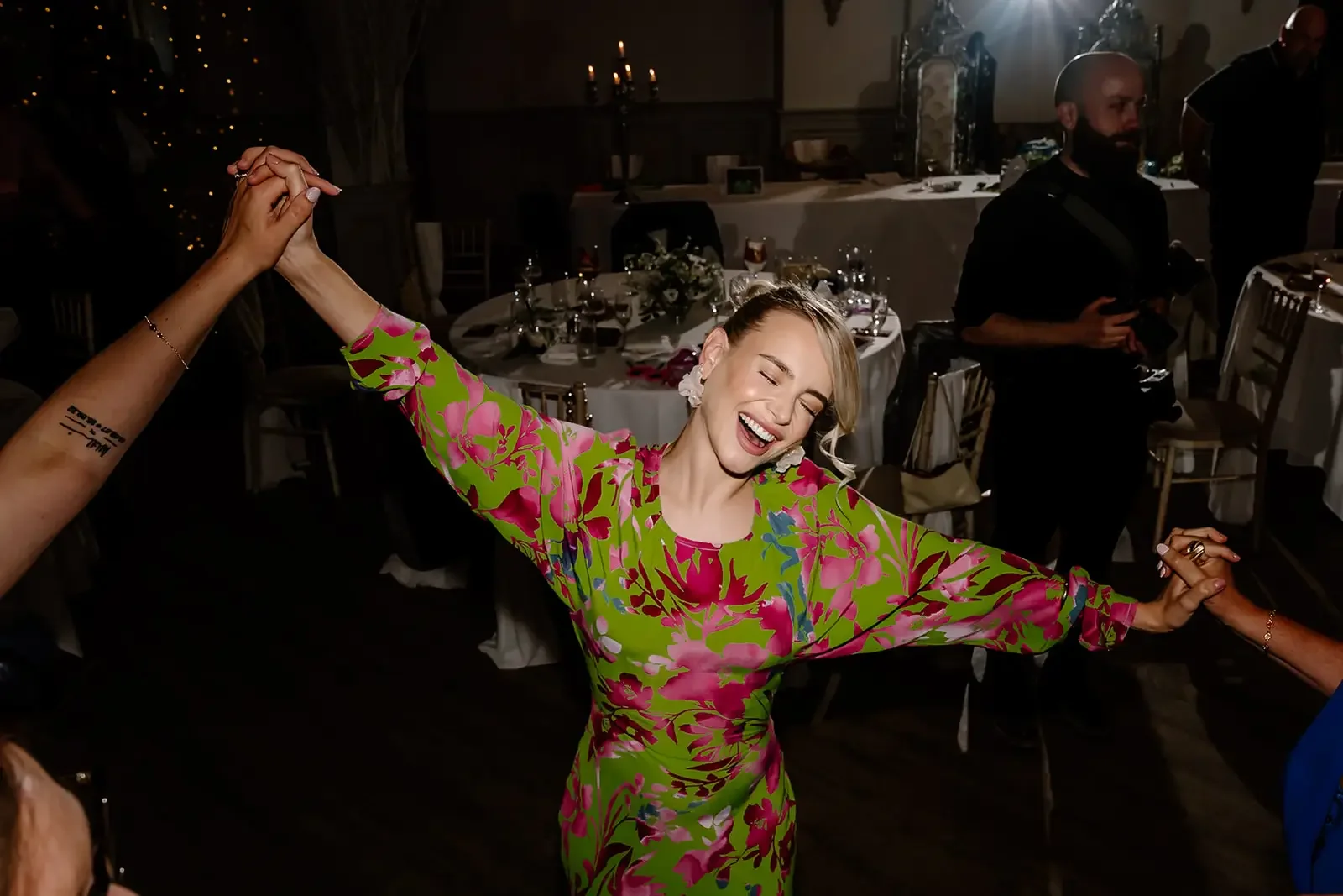A woman with blonde hair in a floral green and pink dress dancing happily at a social event with others holding her hands.