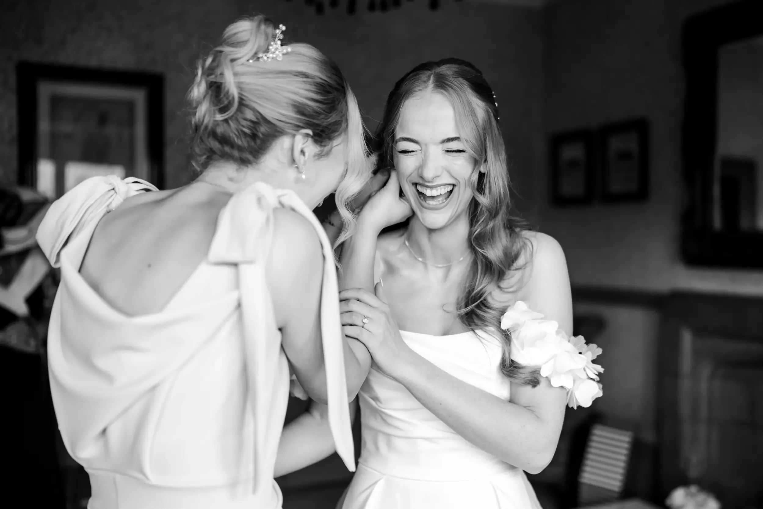 Two women, one possibly a bride, sharing a joyful moment and laughing, in a room with framed pictures on the wall.