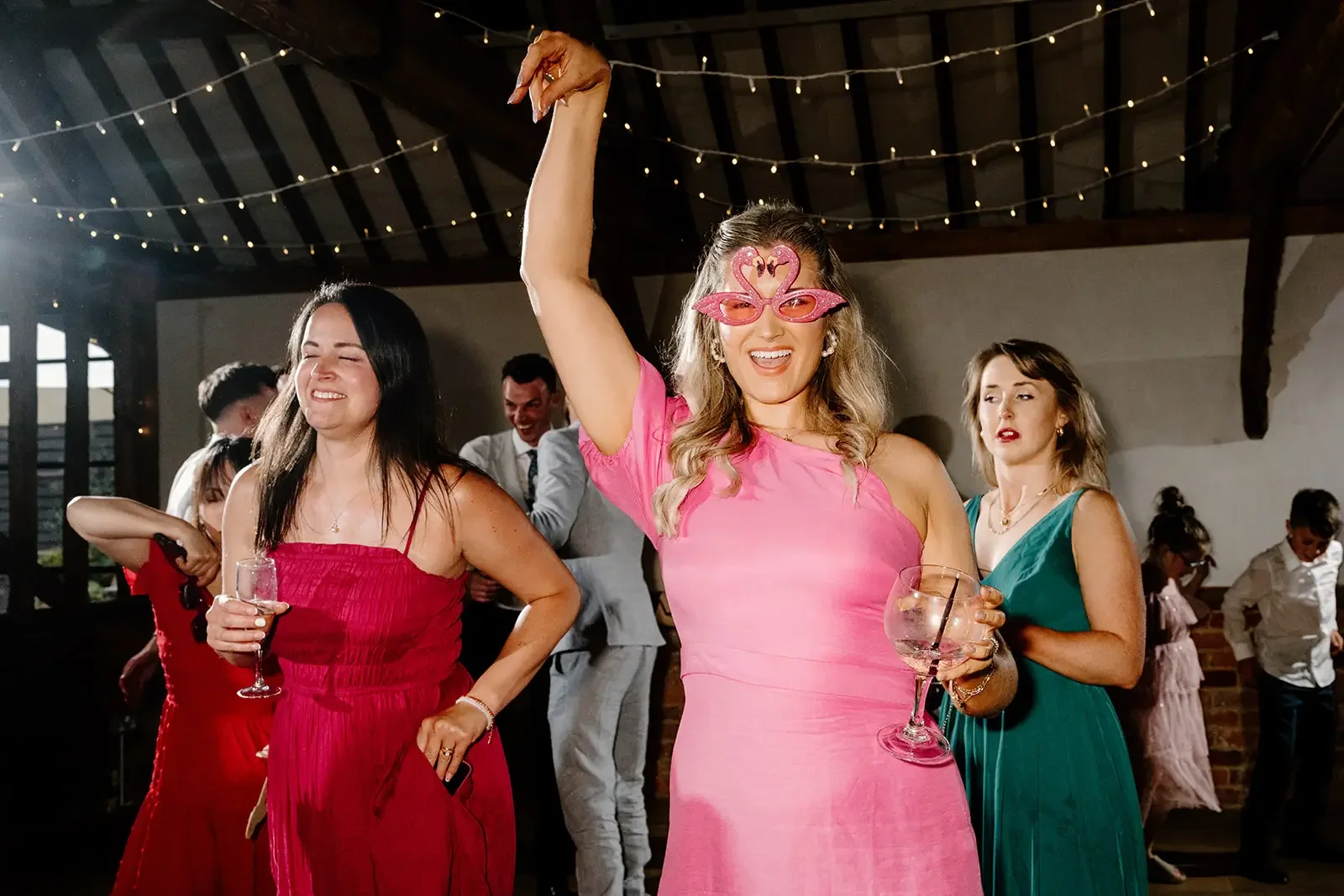 Women dancing and celebrating at a party, one wearing pink sunglasses with flamingos and a pink dress, holding a large cocktail glass, others in colorful dresses, with string lights overhead.