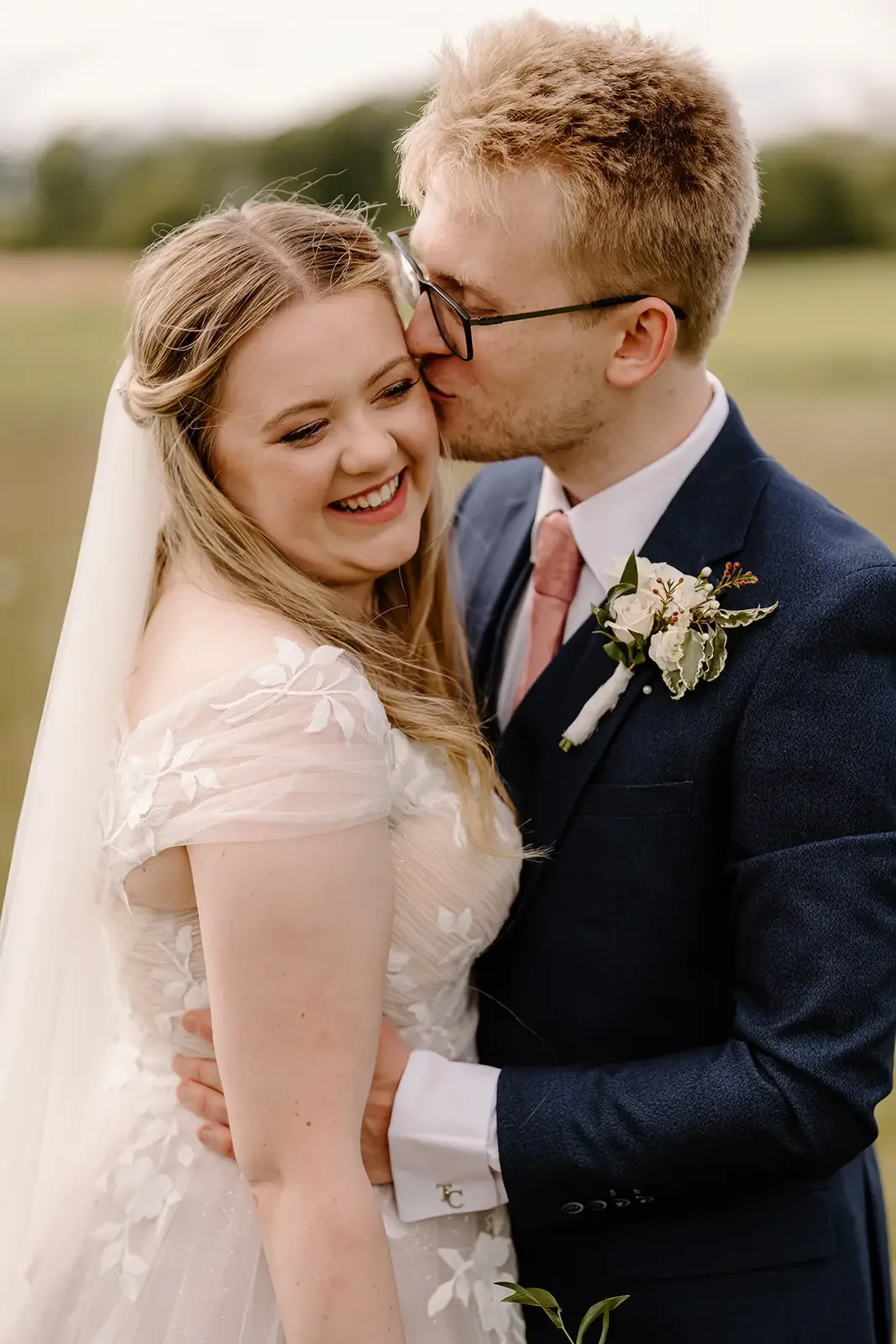 A newlywed couple embracing outdoors, with the groom kissing the bride's temple. The bride is smiling and wearing a white wedding dress with floral embroidery, and the groom is dressed in a navy suit with a pink tie and boutonniere. The background is